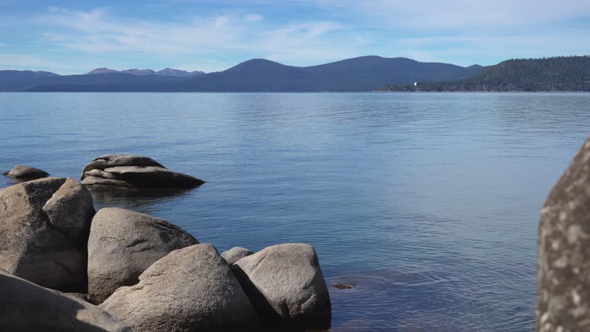 Granite boulders sit along the shoreline of calm Lake Tahoe with distant mountains