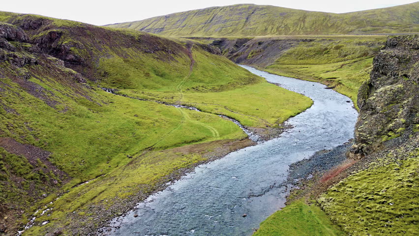 Aerial drone view of a river winding through a narrow canyon surrounded by grassy volcanic hills and rugged cliffs in the Icelandic countryside