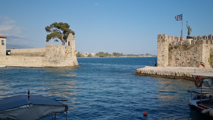 Scenic view of the historic Venetian harbor in Nafpaktos, Greece, with stone castle walls, calm sea, and Greek flag during golden hour