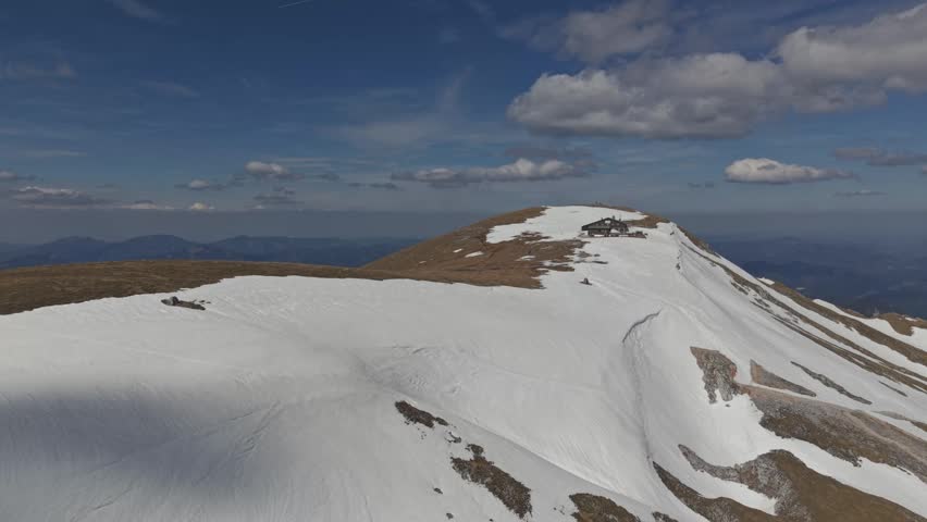 Aerial view of a rugged mountain ridge partially covered with snow, featuring a small alpine hut near the summit.