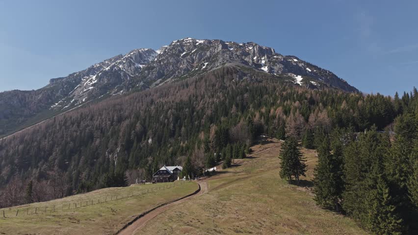 A scenic mountain landscape featuring a rustic cabin nestled among dense forest and rolling hills. Snow-capped peaks rise in the background under a clear blue sky.