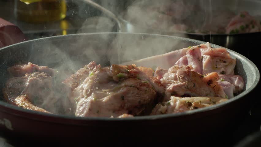 Lamb pieces are cooked in a pan with spices. The meat sizzles and steam rises. Tongs are used to turn the meat to ensure even cooking and flavor.