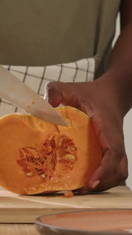 Vertical video: Holding halved squash, home cook in apron scooping seeds, cutting wedges with knife. Pumpkin, kitchen, prep, hands, counter, slice, vertical video