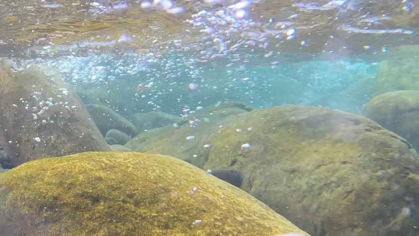 Clear stream flowing through submerged rocks with bubbles underwater at riverbed.