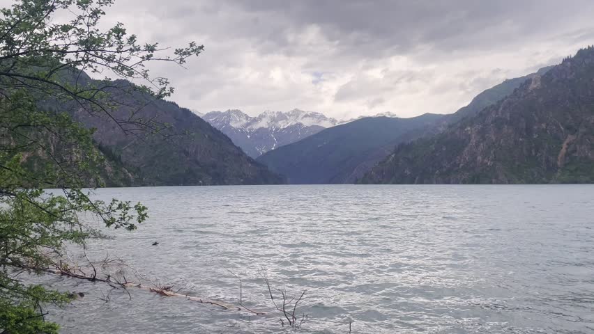 Panoramic view of Sary-Chelek Lake in Kyrgyzstan, featuring pristine alpine waters surrounded by lush green forests and snow-capped mountain peaks under a dramatic cloudy sky in spring