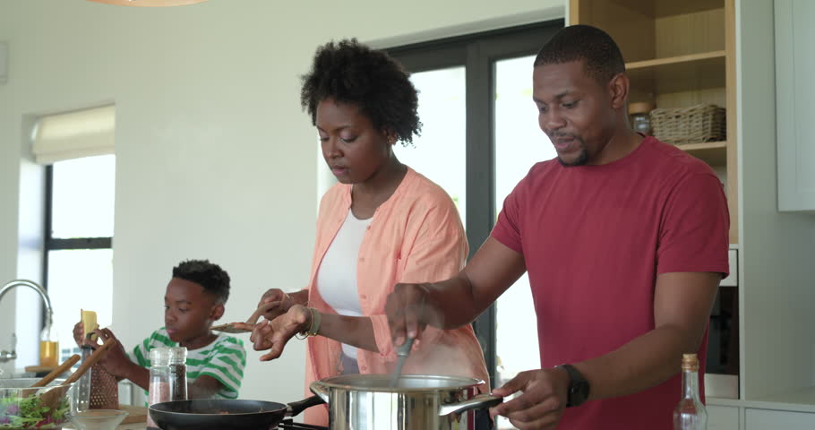 After tasting sauce African American couple stirring pot and boy mixing salad at kitchen island. Family, culinary, modern, vibrant, teamwork, homestyle, domestic