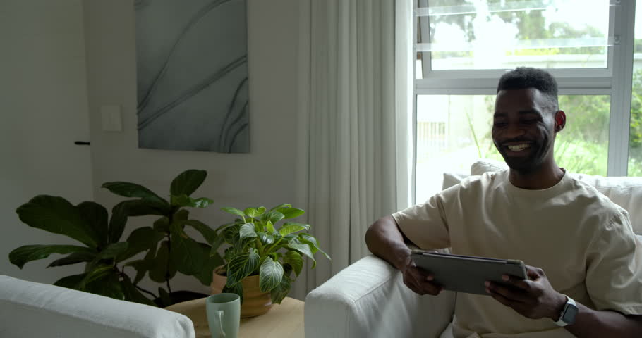 Adult African man smiling and tapping tablet, reacting to entertaining content in armchair at home. Screen, lifestyle, cozy, daylight, window, greenery, smartwatch