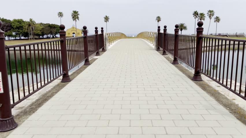 POV shot walking toward a symmetrical pedestrian bridge over water in a quiet public park. Centered view of paved walkway, metal railings, and calm waterfront under cloudy daylight.