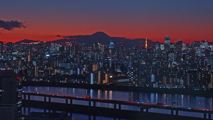 A cinematic zoom shot of the Tokyo metropolis at dusk. The sky is a deep crimson behind the silhouette of Mount Fuji, while city lights and a river create a vibrant urban scene.