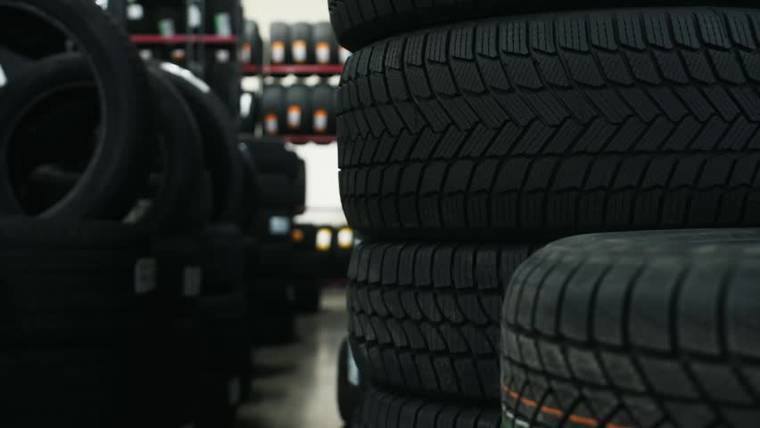 Black rubber tires are stacked in an automotive shop, with additional tires visible on shelves in the background under bright lighting