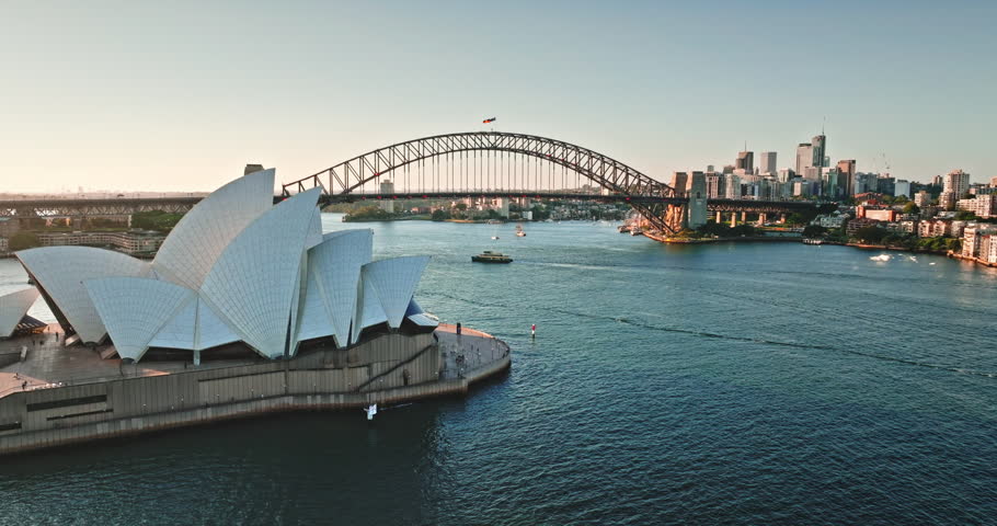 Australia, Sydney cityscape showing the iconic Opera House and Harbor Bridge across the bay at sunset, boats navigating the water and modern city urban buildings in the distance. Aerial view panorama
