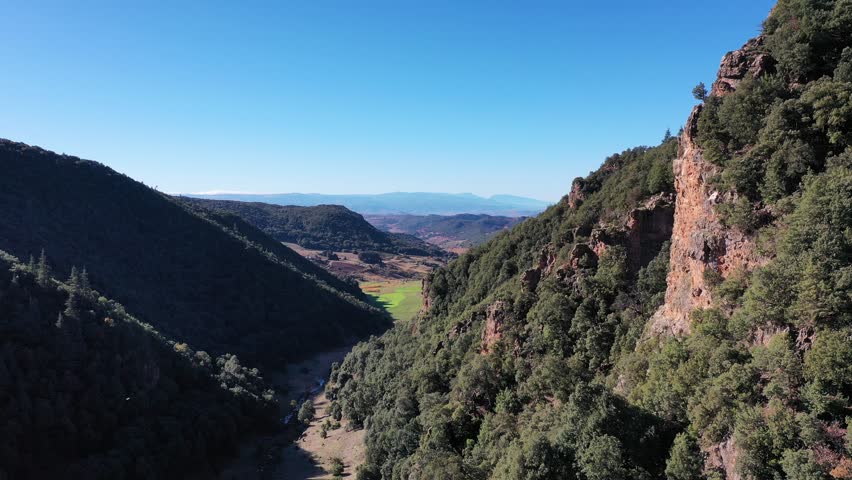 A cinematic 4K drone shot looking down a rugged mountain valley near Khenifra, Morocco; featuring steep limestone cliffs, dense cedar forests, and a distant view of lush green pastures under a clear b