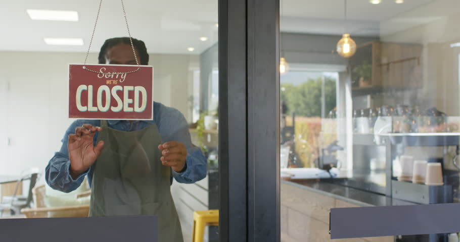 African American male barista opening coffee shop unhooking sign flipping to open peering outside. Cafe, hospitality, welcoming, modern, warm, cozy, customer service