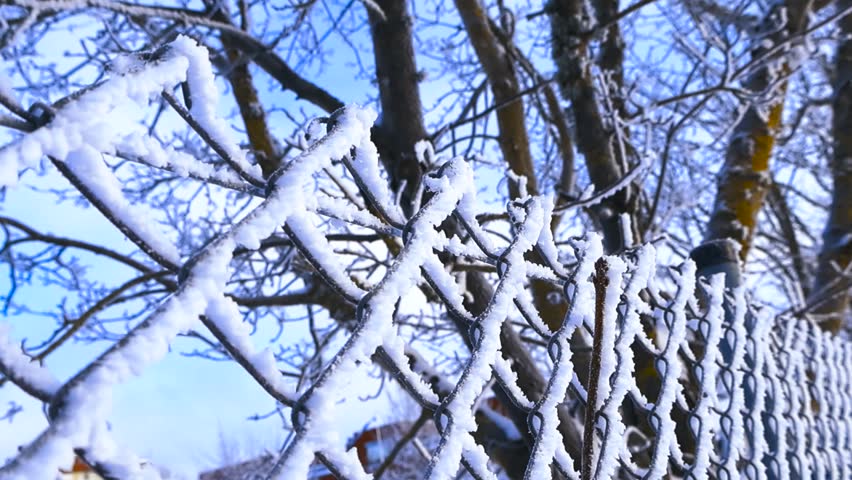 Frosty chain link fence covered with snow and hoarfrost on a cold winter day, with blurred trees and blue sky in the background, showing a beautiful wintery scene in extreme detail