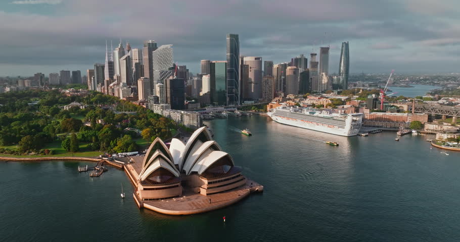 Aerial view of Sydney Opera House at Circular Quay with a cruise ship in the harbor and CBD skyline rising behind, bright summer day over the waterfront and ferry hub. Australia landmarks panorama