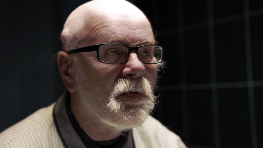 Old man in glasses shaves his beard in front of a mirror in the bathroom