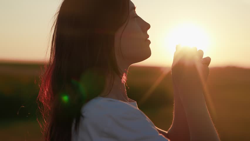 Young girl prays against sunny sky, caring for family children. Faith in God. Close-up Christian woman praying at sunrise. Relaxation meditation in nature. Apology. Hope. Life. Free man. Silhouette.