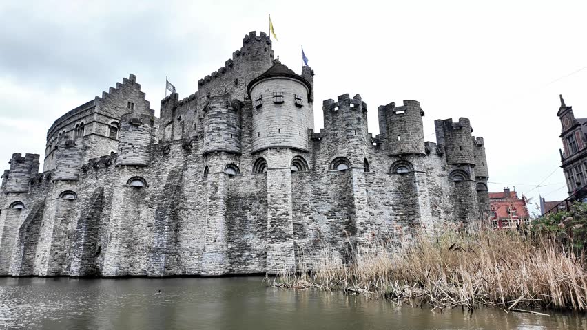 Wide shot of Gravensteen Castle in Ghent from across the water.
This horizontal video captures the imposing Gravensteen (Castle of the Counts) in Ghent. The medieval fortress, built from grey stone with battlements and turrets, rises directly from the water. Tall reeds and dry grass line the foreground at the water
