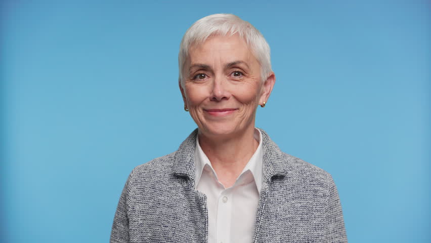Studio portrait of a mature woman with short white hair smiling warmly and making a peace or victory gesture on blue background