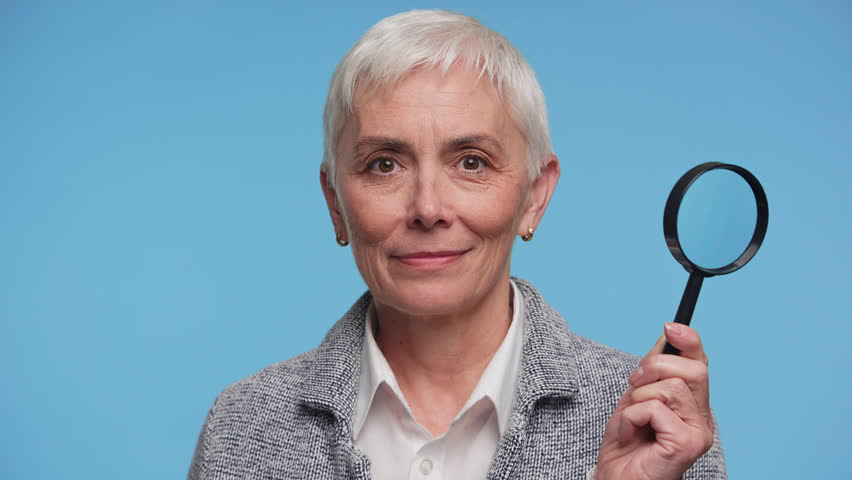 Portrait of an older woman winking and holding a magnifying glass to her eye against a blue background, showing curiosity and close inspection.