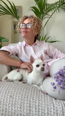 A woman with curly hair and glasses enjoys a peaceful moment at home, gently caressing her cute white Chihuahua on a comfortable sofa. The scene exudes tranquility and companionship.