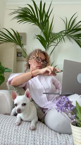 A focused woman uses her laptop on a comfortable sofa, accompanied by her peaceful pet dog. Capturing the essence of remote work and a cozy home environment.