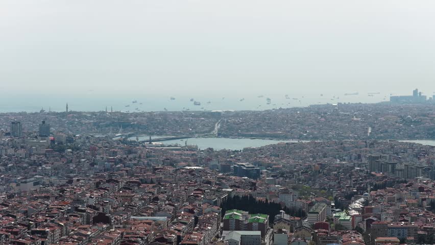 Expansive aerial showing urban landscape of Istanbul with Golden Horn inlet, Galata Bridge, numerous cargo ships on Sea of Marmara under hazy sky