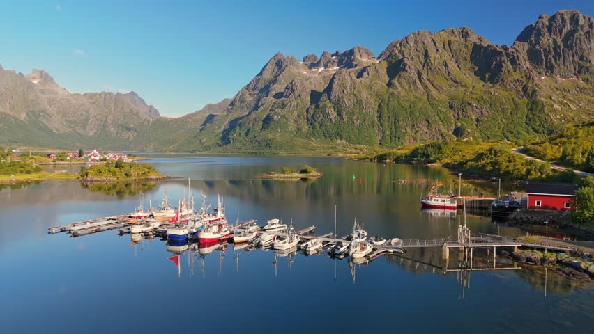  Many boats are docked at the island harbor in the Lofoten Islands, Norway. Drone aerial shot shows sharp mountains reflecting in the calm fjord water of the Lofoten Islands, Norway