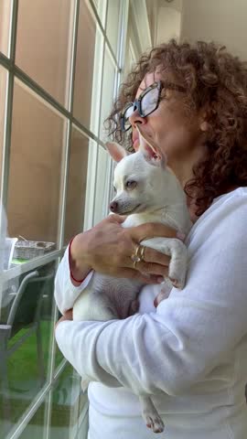 A woman with curly hair and glasses tenderly holds her small white Chihuahua dog while looking out a large window, creating a domestic scene of calm and affection. The cute pet yawns softly in her arms, showcasing their loving bond.