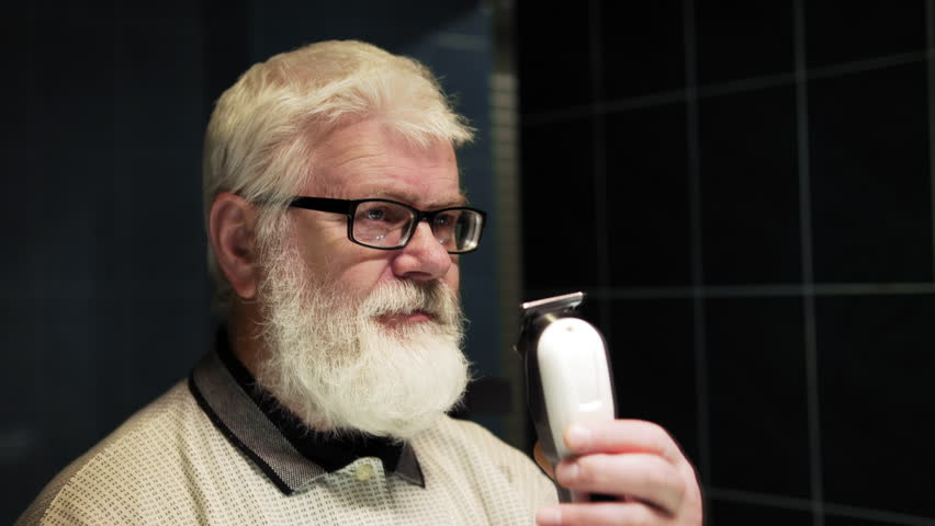 Pension man in glasses cutting his own hair with a trimmer in front of a mirror