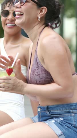 Vertical video: Laughing woman in purple bikini offering cocktail to friend at patio, chat starting. Poolside, outdoor, terrace, drink, straw, strawberry, vertical video