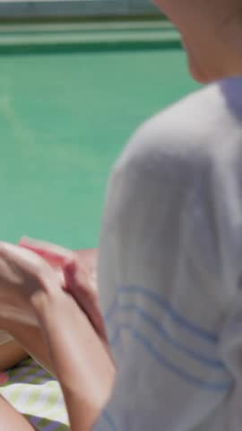 Vertical video: Copying girl in pink rash top tugging white shades after adult patting legs at pool. Sunny, outdoor, backyard, sunglasses, sunscreen, lotion, vertical video