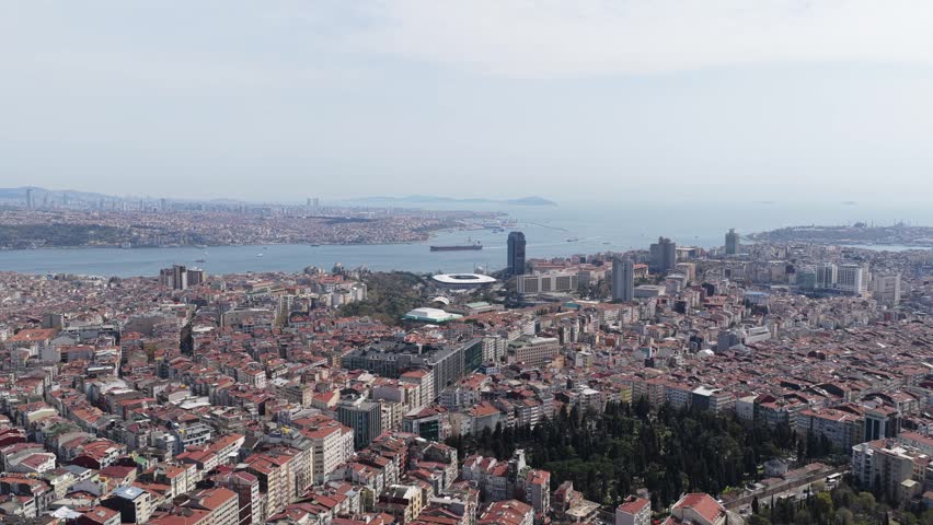 Extensive aerial panorama of Istanbul European side, featuring dense urban landscape of red roofed buildings, modern skyscrapers, expansive Bosphorus strait under hazy sky