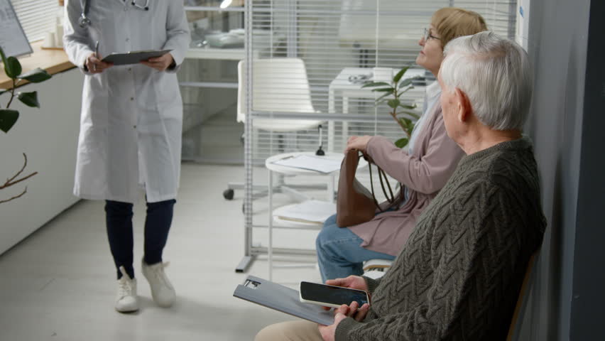 Elderly man following female doctor to consultation room while another patient waiting in queue at modern clinic