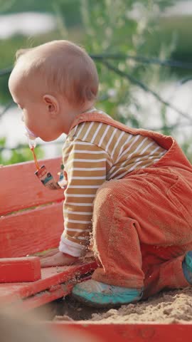 A small baby is climbing on a red bench. The baby wears an orange outfit with stripes and is surrounded by greenery near a lake.
