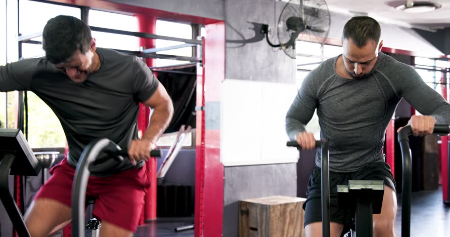 Diverse male friends mounting adjacent air bikes at gym, pedaling hard, ending with high-five. Fitness, teamwork, motivation, camaraderie, training, exercise, athleticism