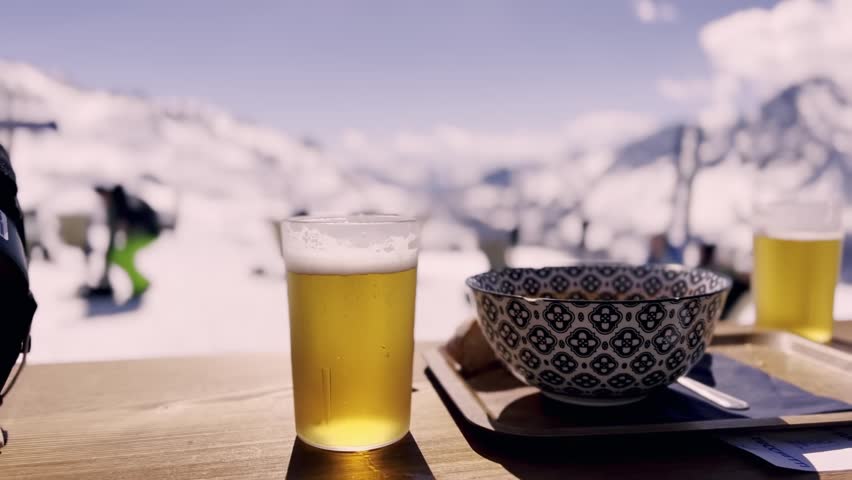 Refreshing glass of beer and a warm bowl of soup resting on a wooden table at a ski resort, with skiers and snowy mountain peaks creating a beautiful blurred background on a sunny day