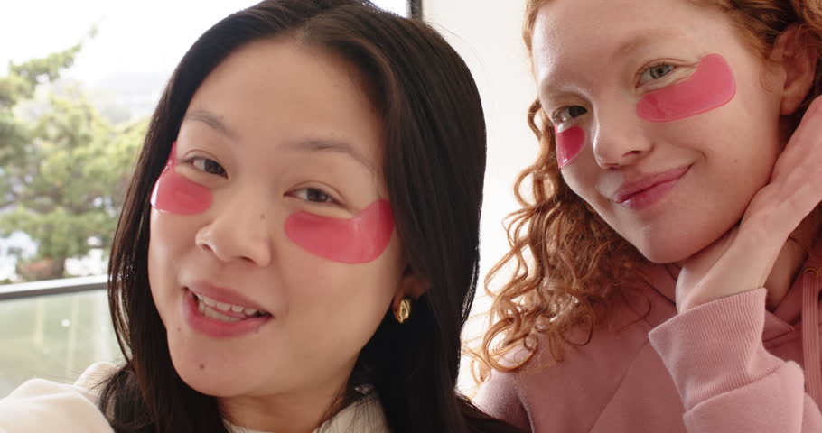 Diverse female friends filming selfie at window wearing pink under-eye patches, sharing skincare. Companionship, friendship, casual, home, balcony, natural, light