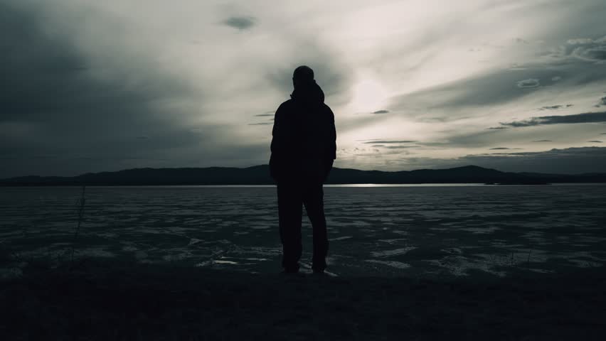 A black and white silhouette of a lonely man standing alone on the shore of an icy lake. The psychological state of a person.