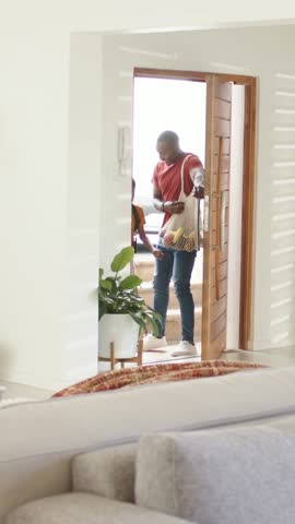 Vertical video: Opening door man in red tee holding grocery bag and letting family enter at home. Entrance, trio, livingroom, daylight, threshold, plant, vertical video