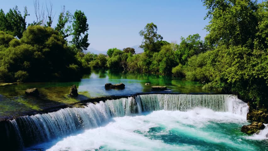 View of Manavgat Waterfall from above in the National Park on a sunny summer day. Unique azure watercolor color with splashes. A wide stream of water falls from a low height. Antalya, Dron, 4K