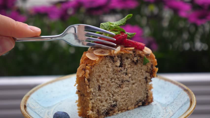 Close up view of a person using a fork to cut a delectable slice of homemade cake, garnished with fresh mint leaves, sliced strawberries, almonds, and a single blueberry on the side