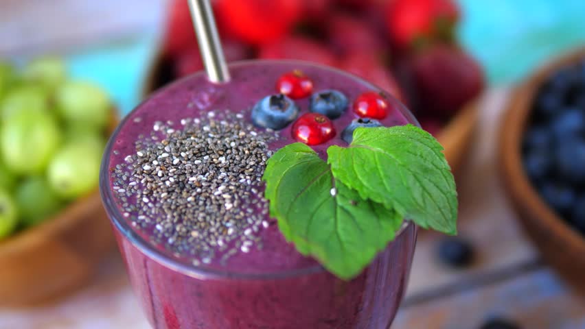 Close up of a healthy blended berry smoothie garnished with chia seeds, blueberries, red currants, and mint, slowly rotating with fresh fruits in the background for a nutritious vegan breakfast