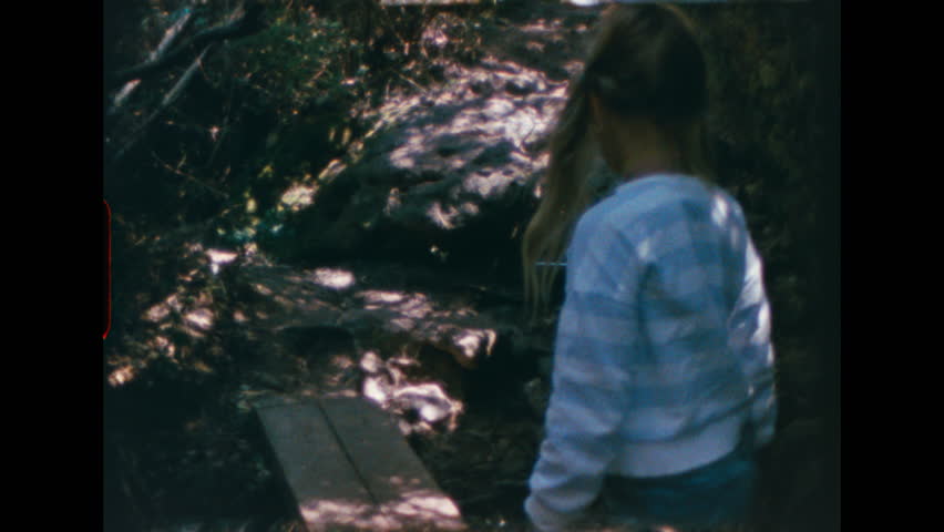 Girl in white shirt and jeans climbing rocky forest trail, stumbles but keeps balance, shot on super 8 film with retro grain and color