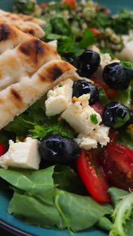 Delicious and healthy greek salad served on a green plate, featuring fresh lettuce, feta cheese, olives, and tomatoes with pita bread, hummus, and a spicy dip on the side for a complete meal