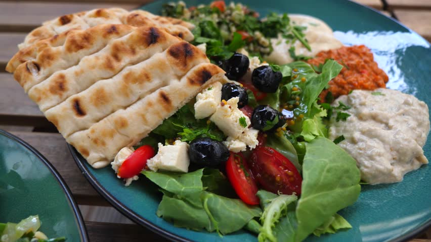 Mediterranean mezze spread with fresh salad topped with feta and olives, pita bread, hummus, baba ghanoush and other dips served on a rustic wooden table for sharing