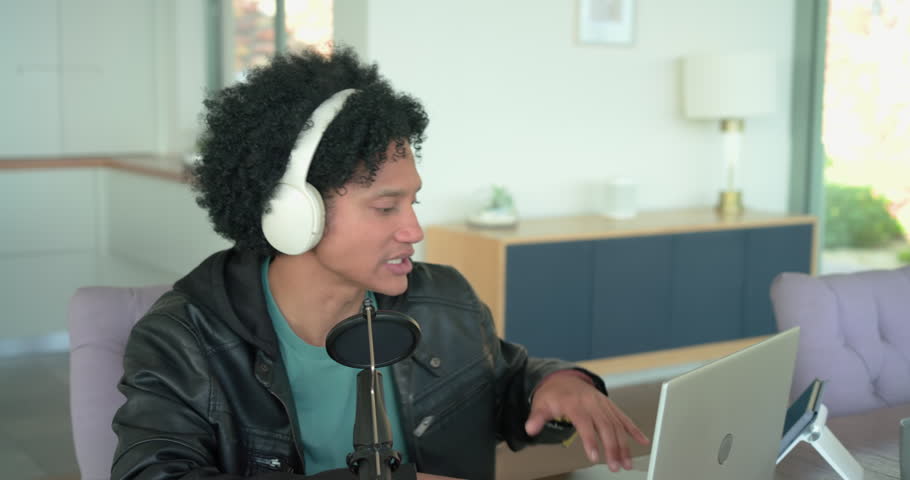 Man in his thirties entering office, donning headphones, using laptop and mic to record, copy space. Digital creator, content production, modern workspace, sound recording, remote work