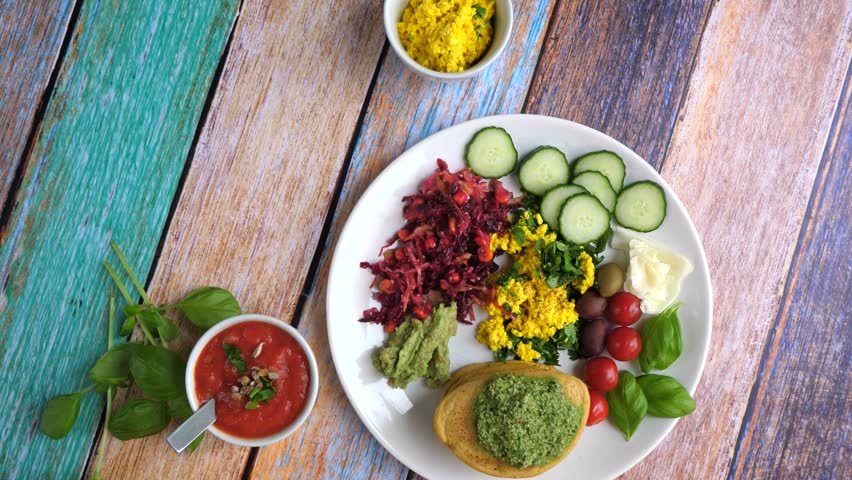 Top view of a person serving a colorful and healthy vegan brunch with pancakes, pesto, tofu scramble, fresh vegetables, and dipping sauces on a vibrant wooden table for two people