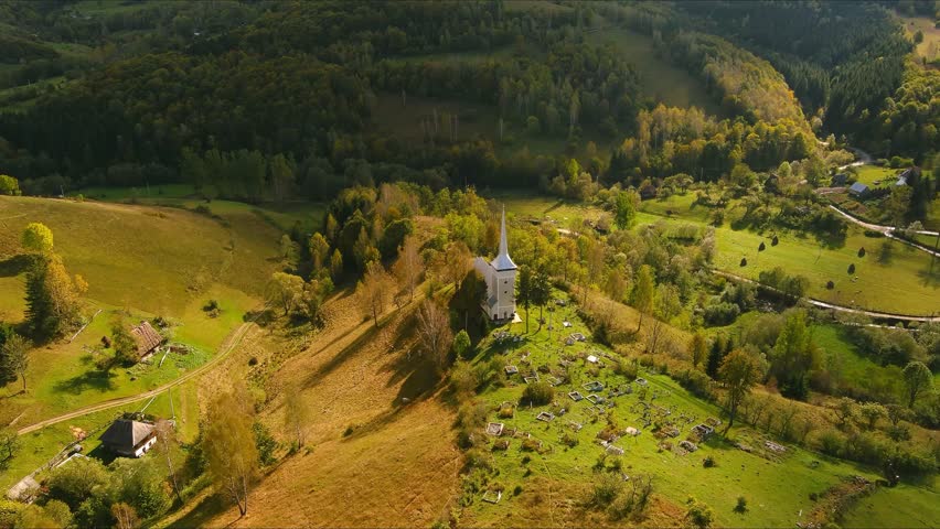 Aerial view of a white church with a tall spire on a grassy hill surrounded by a cemetery and autumn trees in Salciua de Jos, Alba County, Romania.