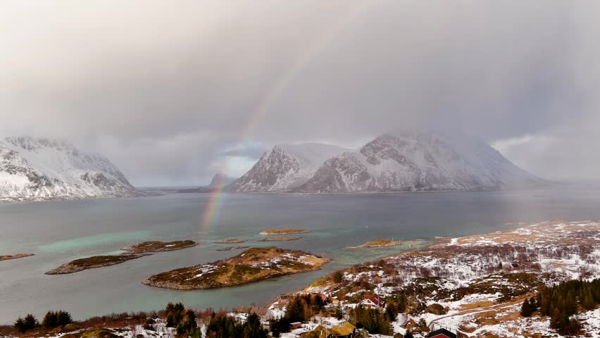 Aerial view of a vibrant rainbow stretching over snow-capped mountains and small islands in a calm fjord under a cloudy sky in Nordland, Norway.